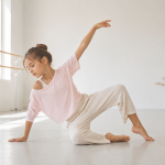 una niña practicando danza contemporanea vestida con una camiseta rosa y pantalónes blancos. Está sentada en el suelo con un brazo hacia arriba y la mirada proyectada hacia abajo.