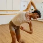 un chico practicando un movimiento de danza contemporánea. Está en un aula y se ve una barra de ballet al fondo. Él va vestido en tonos claros.